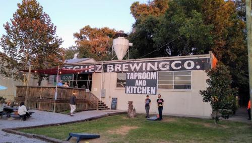 Exterior of a brewery with a large sign reading "Taproom and Kitchen," surrounded by trees and outdoor seating.