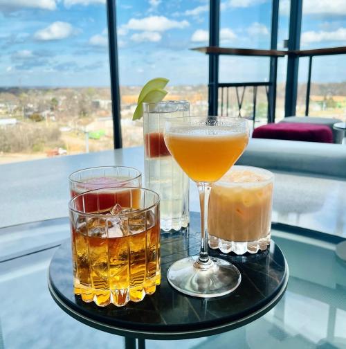 A selection of four colorful cocktails on a round table with a scenic view of the sky and landscape in the background.