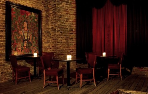 Dimly lit interior of a rustic bar with wooden floors, red curtains, and a large framed painting on the brick wall.