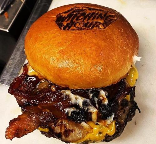 A delicious burger with a toasted bun, melted cheese, crispy bacon, and grilled onions, branded with a logo.