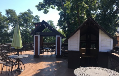 Outdoor deck with tables, chairs, and small structures, surrounded by trees and string lights.
