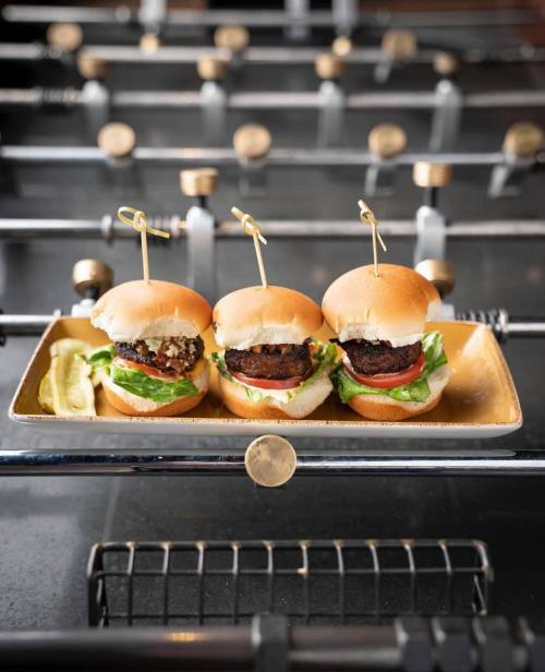 Three mini burgers on a tray, each with lettuce, tomato, and a skewer, served with a pickle on the side.