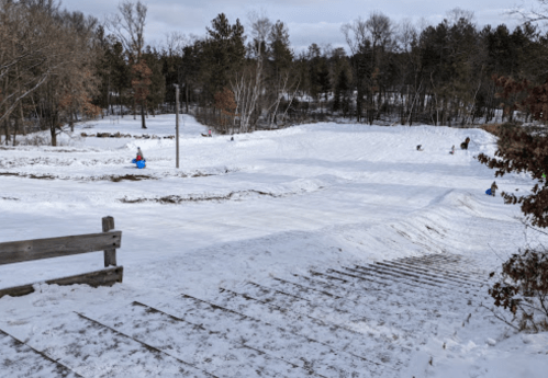 A snowy landscape with a sledding hill, trees in the background, and people enjoying winter activities.
