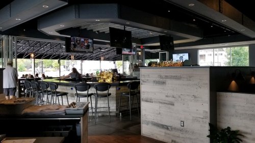 Modern bar interior with high stools, multiple TVs, and a well-stocked counter, featuring a bright, open atmosphere.