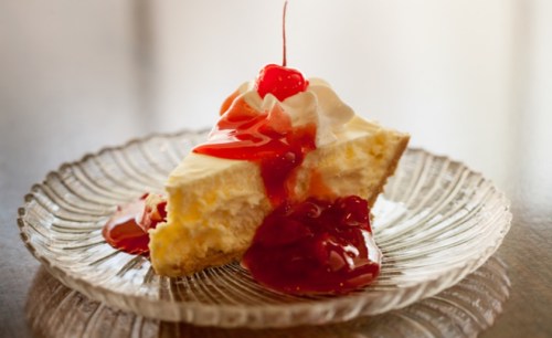 A slice of cheesecake topped with cherry sauce and whipped cream on a decorative glass plate.