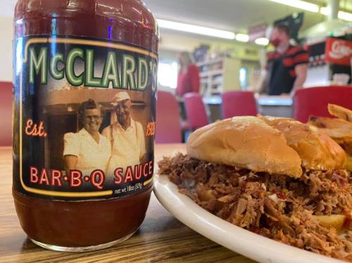 A bottle of McClard's BBQ sauce next to a plate of pulled pork sandwich and fries in a diner setting.
