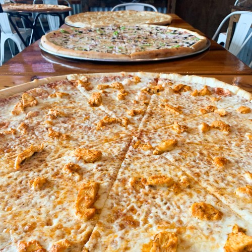 A close-up of a cheese pizza with chicken pieces, with two large pizzas in the background on a wooden table.
