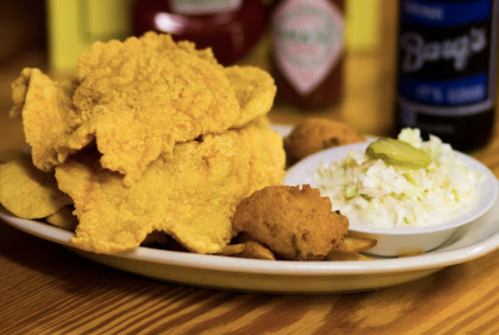 A plate of fried fish, coleslaw, hushpuppies, and pickles, with condiments in the background.