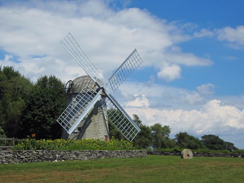 A traditional windmill stands beside a field, surrounded by greenery and under a blue sky with fluffy clouds.