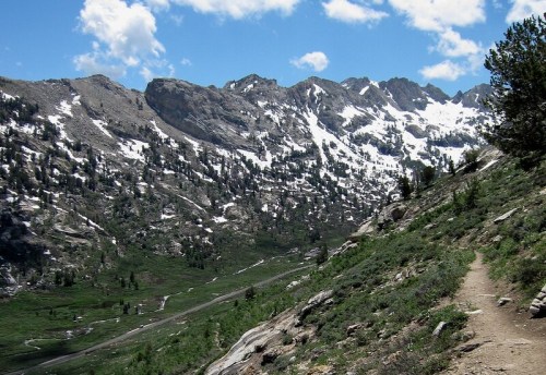 A scenic mountain landscape with snow-capped peaks, green valleys, and a winding dirt path under a blue sky.