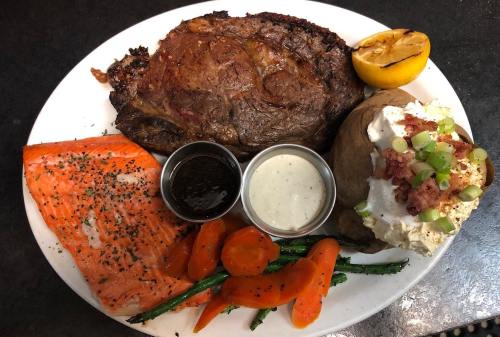 A plate featuring a ribeye steak, salmon, baked potato with toppings, carrots, green beans, and a lemon wedge.