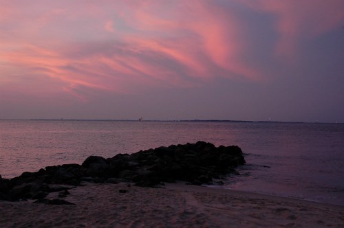 A serene beach scene at dusk, featuring a calm sea, rocky shoreline, and a colorful pink and purple sky.