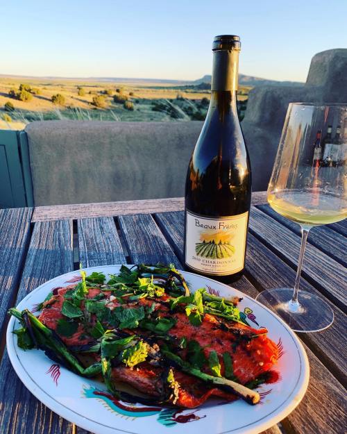 A plate of grilled salmon topped with herbs, a bottle of Chardonnay, and a glass of wine on a wooden table outdoors.