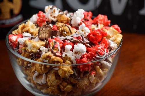 A clear bowl filled with colorful popcorn, including red, white, and caramel varieties, topped with chocolate pieces.