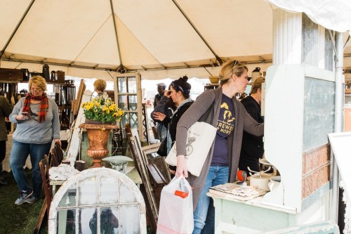 A bustling market tent filled with people browsing vintage items and decor, with plants and furniture on display.