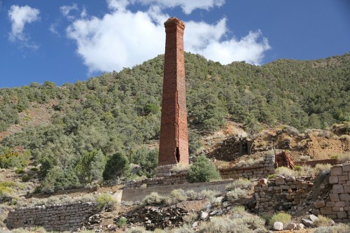 A tall, red brick chimney stands among green hills and blue skies, remnants of an old structure in a rugged landscape.
