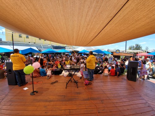 A lively outdoor event with a large crowd gathered under a shaded area, enjoying music and activities.