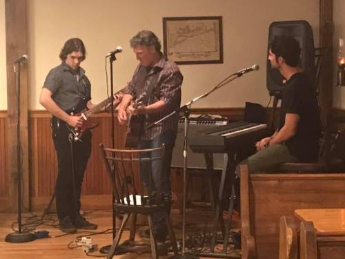 Three musicians perform on stage: one on guitar, one on keyboard, and another playing electric guitar in a cozy venue.
