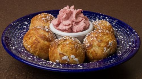 A blue plate with five golden-brown pastries dusted with powdered sugar, served with a bowl of pink whipped topping.