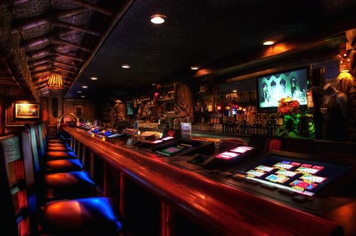 A dimly lit tiki bar with a long wooden counter, colorful lights, and a variety of drinks displayed on the shelves.