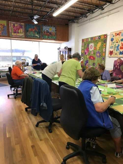 A group of people working on a craft project at a long table in a bright, spacious room. Colorful artwork decorates the walls.