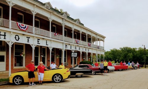 A group of people in colorful shirts stands beside classic cars in front of a historic hotel decorated with bunting.