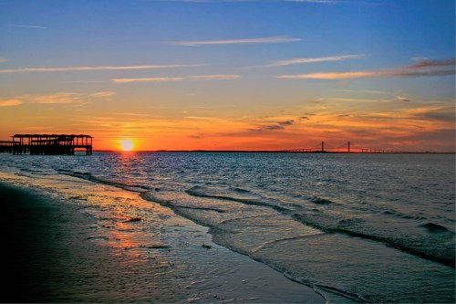 A serene sunset over the water, with gentle waves and a distant bridge silhouetted against the colorful sky.