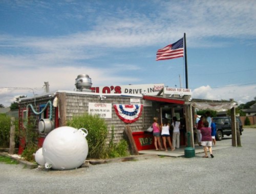 A small drive-in restaurant with a flag, people entering, and a rustic exterior surrounded by greenery.
