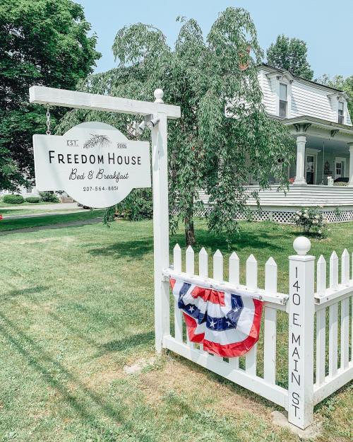 Sign for Freedom House Bed & Breakfast with a white picket fence and patriotic bunting, set in a green landscape.