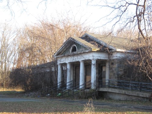 An abandoned building with columns, overgrown vegetation, and a weathered facade, set in a wooded area.