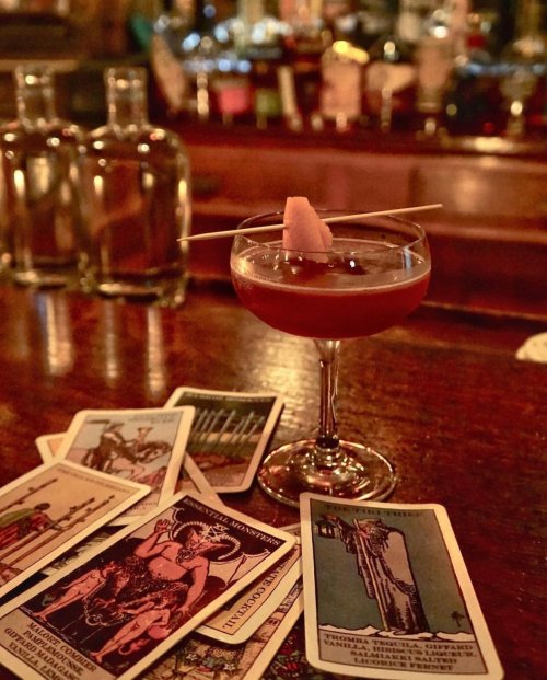A cocktail in a coupe glass garnished with a fruit slice, surrounded by tarot cards on a wooden bar.