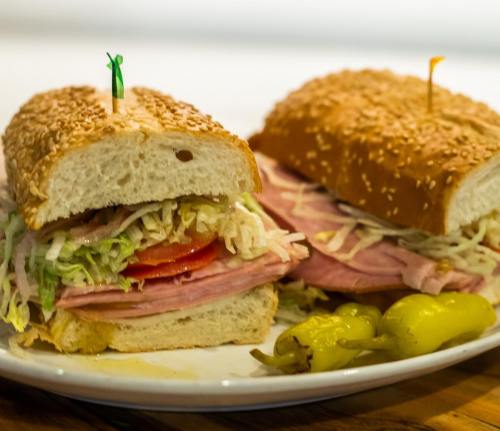 A close-up of a sesame seed sandwich filled with ham, lettuce, tomato, and peppers, served on a white plate.