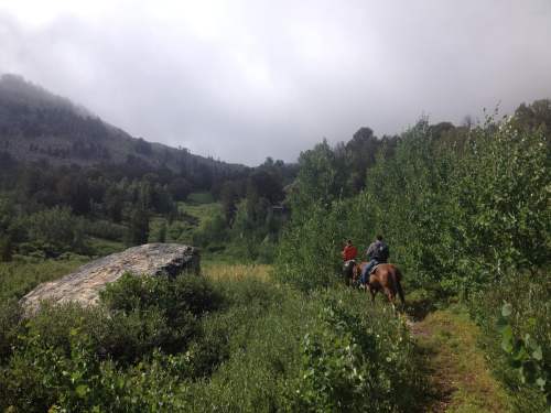 Two people on horseback ride through a lush, green landscape with a rocky outcrop and misty mountains in the background.