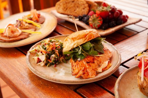 A grilled salmon sandwich with greens on a plate, accompanied by a colorful salad and assorted fruits in the background.