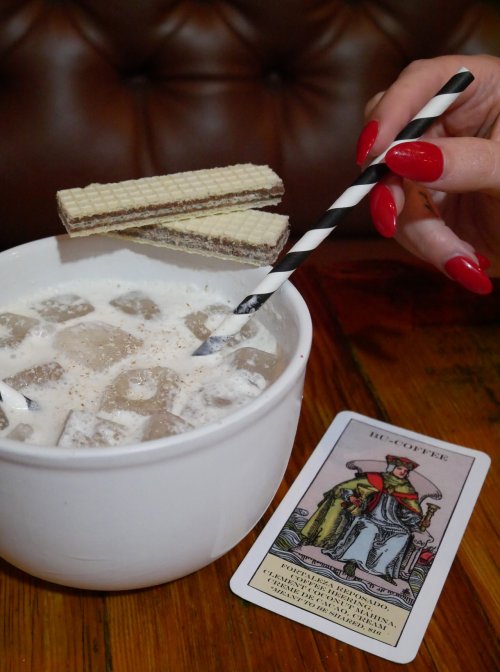 A hand with red nails holds a straw in a white cup of iced coffee, accompanied by wafer snacks and a tarot card.