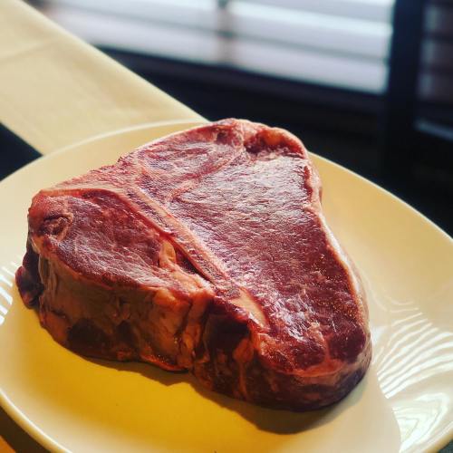 A large, raw T-bone steak on a white plate, resting on a table with a blurred background.