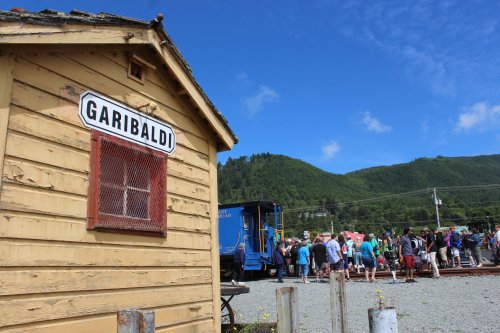 A wooden building labeled "Garibaldi" with a crowd gathered nearby, set against a backdrop of green hills and blue sky.