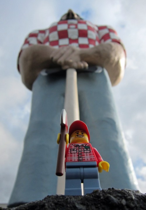 A LEGO figure in a red checkered shirt stands in front of a giant statue of a lumberjack holding an axe.