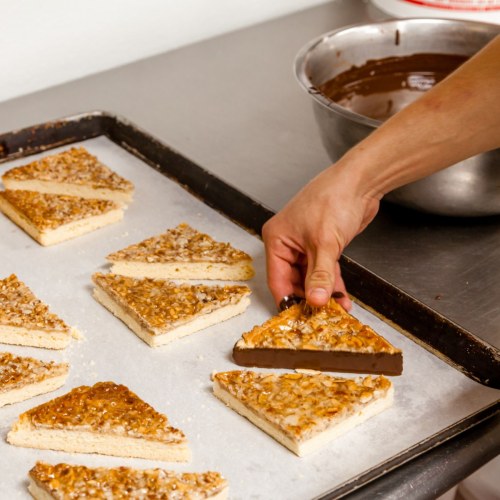 A hand lifts a chocolate-coated treat from a tray of triangular pastries topped with caramelized nuts.