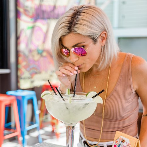 A woman with blonde hair and sunglasses sips a large margarita with multiple straws at a vibrant outdoor setting.