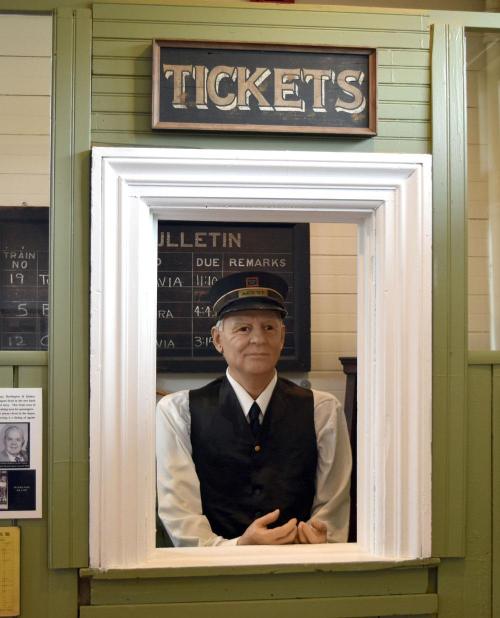 A lifelike mannequin in a ticket booth, wearing a uniform and cap, with a sign reading "TICKETS" above.