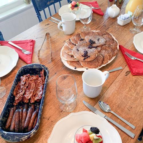 A breakfast table with pancakes, bacon, fruit, and drinks, set with red napkins and glassware.