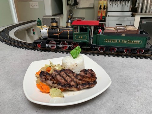 A plate of grilled steak with rice and vegetables, with a model train in the background.