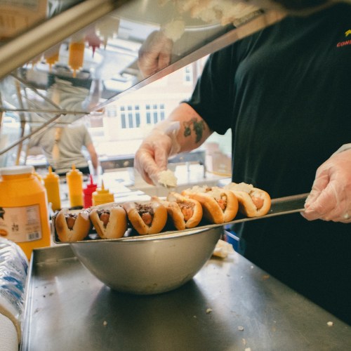 A person in gloves prepares hot dogs in a kitchen, with condiments visible in the background.