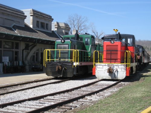 Two vintage locomotives, one green and one red, parked side by side on a railway track near a historic station.