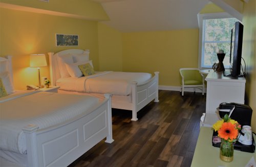 Brightly lit room with two white beds, a TV, and a small seating area, featuring yellow walls and wooden flooring.