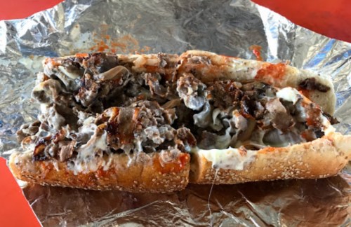 A close-up of a loaded sandwich with beef, melted cheese, and sauce, wrapped in foil on a red surface.
