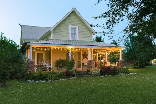 A charming green house with a porch adorned with string lights, surrounded by lush greenery and a well-kept lawn.