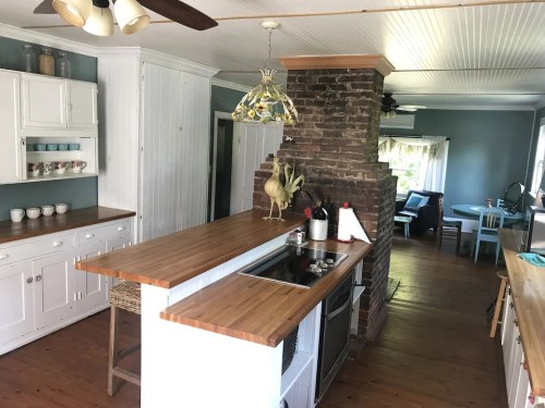 Bright kitchen with wooden countertops, white cabinets, a brick column, and a dining area in the background.