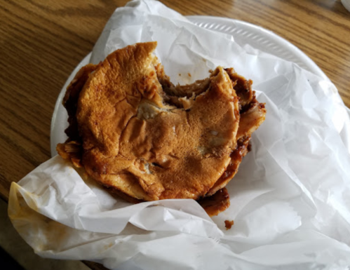 A partially eaten sandwich on a paper plate, wrapped in white paper, resting on a wooden table.